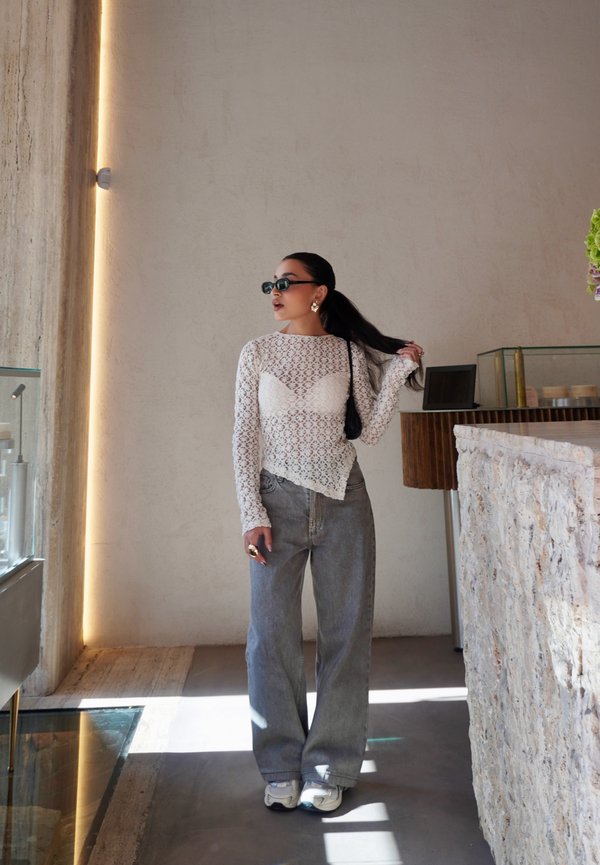 Oberteil aus weißer Spitze mit langen Ärmeln und transparentem Design, kombiniert mit hellblauen, hoch taillierten Jeans und weißen Sneakers. Im Hintergrund ist eine steinerne Arbeitsplatte zu sehen.