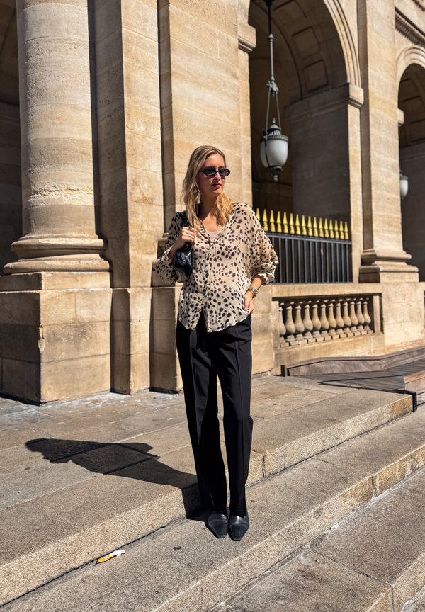 Woman wearing sunglasses and leopard print blouse stands on stone steps outside a building with large columns and iron railing.
