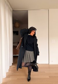 Femme portant un béret noir, un pull bleu marine, une jupe plissée grise, un manteau noir et des bottes, debout à l'intérieur, levant une jambe et souriant avec un sac à main sur l'épaule.
