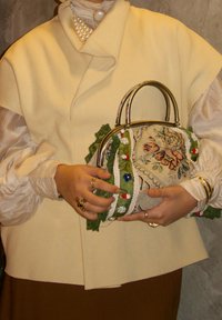 Mains tenant un sac à main brodé de fleurs avec une bordure en dentelle verte, portant un haut crème, un collier de perles, des bagues en or et des bracelets en or.