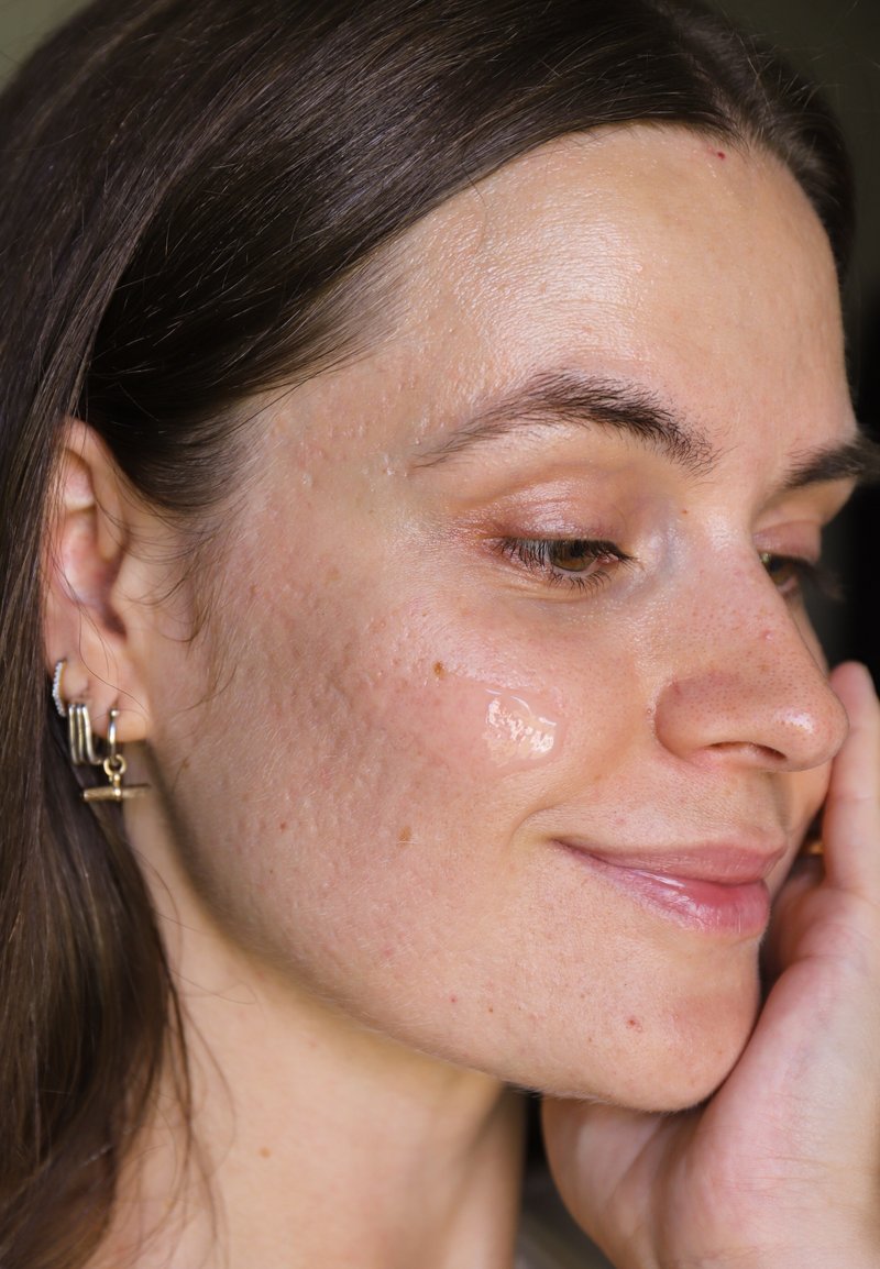 Jeune femme aux cheveux bruns et portant plusieurs boucles d'oreilles touche doucement sa joue sur laquelle un produit de soin de la peau transparent a été appliqué.