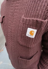 Gros plan sur la poche texturée d'une veste en maille mauve avec un patch logo Carhartt cousu sur la partie supérieure de la poche.