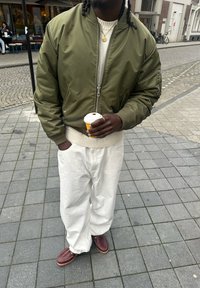 Personne portant une veste vert olive, un pull crème, un pantalon blanc et des chaussures marron, tenant une tasse de café jaune sur un trottoir en ville.