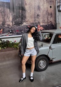 Harmaa bleiseri valkoisen tank topin, harmaiden shortsien, valkoisten nilkkasukkien ja mustien loaferien päällä. Taustalla vintage-auto, jonka pinta on kulunut.