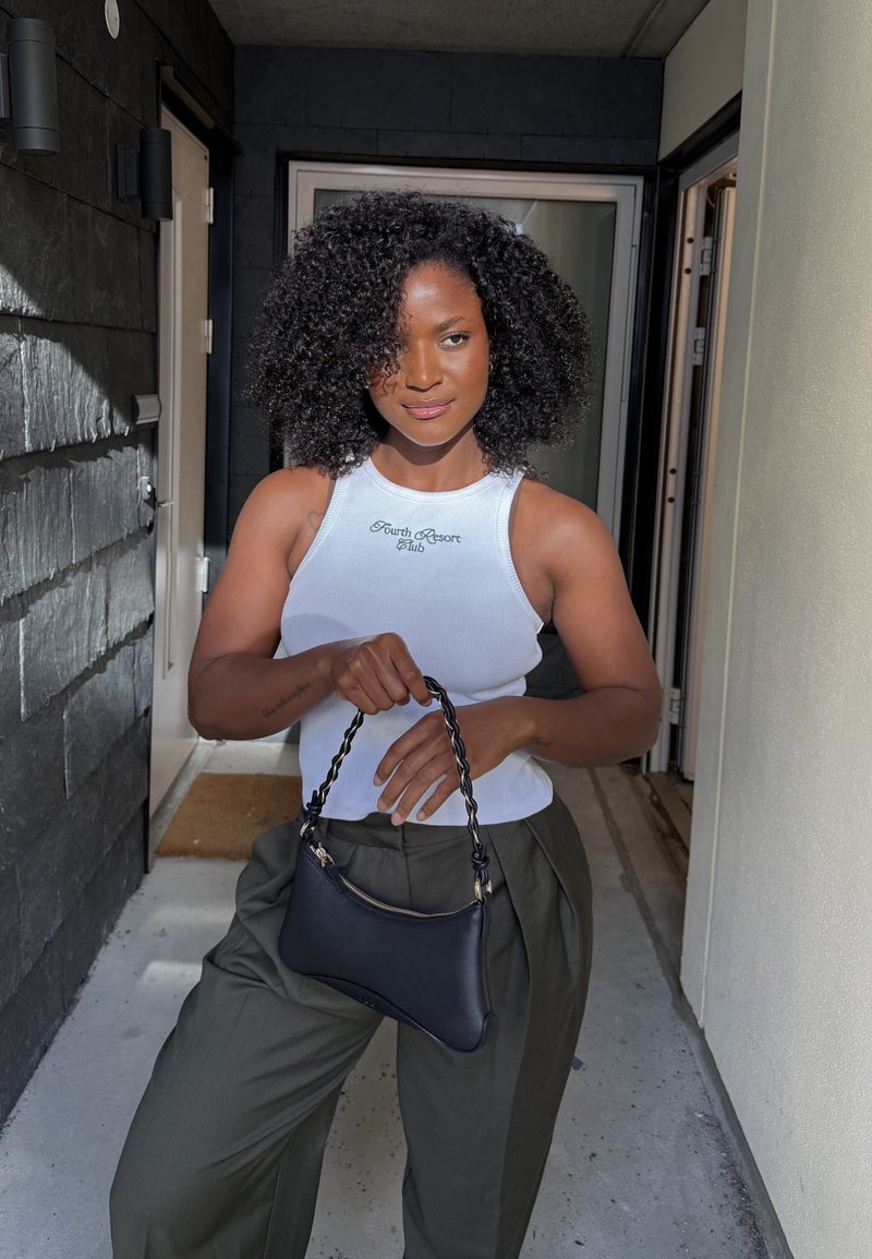 Femme aux cheveux bouclés portant un haut blanc sans manches et un pantalon vert, tenant un petit sac à main noir avec une anse tressée.