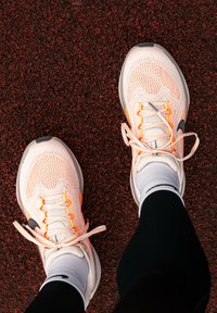 Lätta löparskor i vitt mesh med orange detaljer, utrustade med en vadderad sula, snören och sexkantigt mönster på yttersulan.