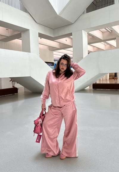 Chándal de satén rosa con rayas blancas, pantalones de pierna ancha y chaqueta con cremallera; combinado con un bolso rosa y zapatillas a juego. Textura suave.