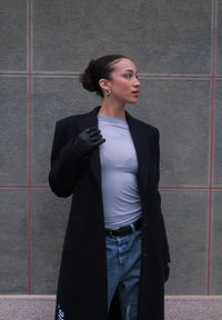 Femme aux cheveux attachés en chignon, portant des boucles d'oreilles créoles, un manteau noir, un haut gris, un jean bleu et des gants noirs, se tenant contre un mur carrelé gris, regardant vers la droite.