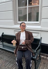 Brun oversized blazer, hvit grafisk t-skjorte, svarte joggebukser med hvite striper, liten brun håndveske, iskald kaffe i klar kopp.