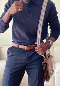 Mann trägt einen dunkelblauen Pullover und Hosen mit braunem Gürtel, hält eine braune Ledertasche mit gestreiftem Schultergurt und zeigt Ringe und Armbanduhr.