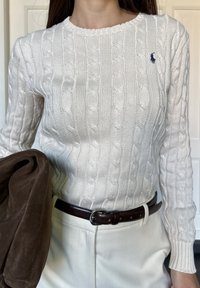 Biały sweter w splot kabelkowy z żebrowanym okrągłym dekoltem i długimi rękawami, zestawiony z brązową zamszową kurtką zarzuconą na ramię oraz białymi spodniami.