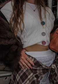 Bijeli pleteni kardigan s šarenim gumbima, slojevit ispod smeđeg kaputa od umjetnog krzna. Sive trenirke i karirana košulja vezana oko struka.