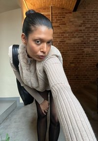 Béžový, texturovaný svetr s vysokým límcem a huňatým předním dílem, s žebrovanými rukávy, spárovaný s průhlednými černými punčochami. Černá taška přehozená přes rameno.