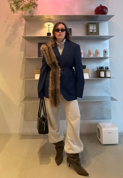 Mujer con blazer azul marino grande y bufanda de piel, pantalones deportivos grises, botas negras, gafas de sol, sosteniendo un bolso negro, de pie en el interior junto a estanterías.