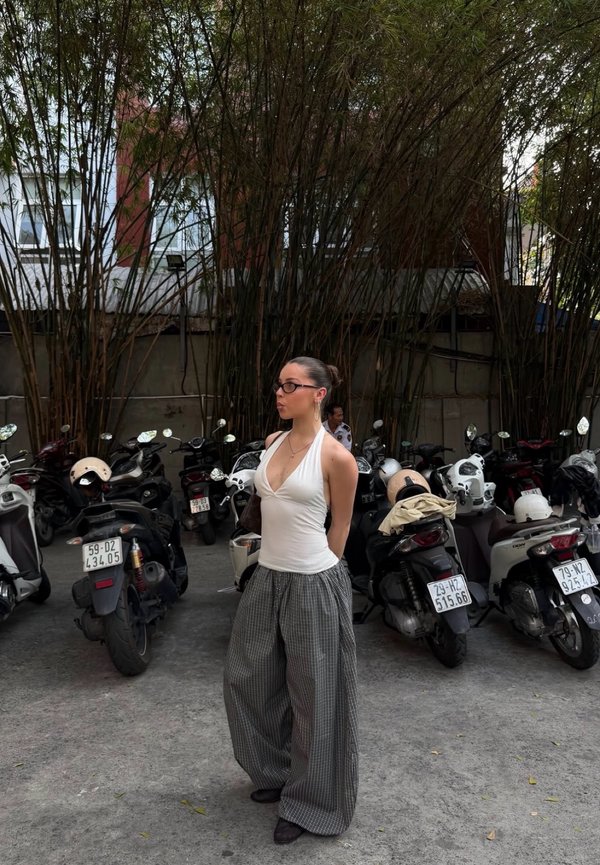 Femme en haut à col licou blanc et pantalon large à carreaux se tient les mains derrière le dos devant des scooters garés et de grandes plantes de bambou.