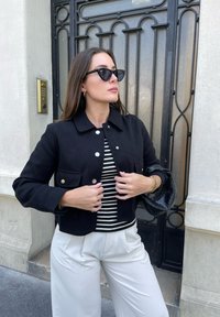 Černá zkrácená bunda se stříbrnými detaily a dvěma předními kapsami, nošená přes černobílé pruhované tričko, doplněná krémovými kalhotami.