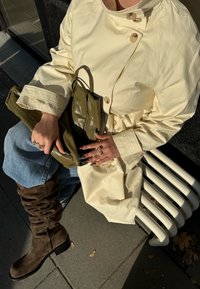 Krém színű trench kabát nagy gombokkal, olívazöld fényes kézitáska, bővülő világoskék farmernadrág és barna, lazán álló térdig érő csizma.