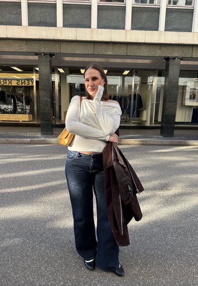 Mujer sonriendo al aire libre con camiseta blanca de manga larga y vaqueros oscuros, sosteniendo una chaqueta marrón y un bolso amarillo, de pie en una calle frente a una tienda.