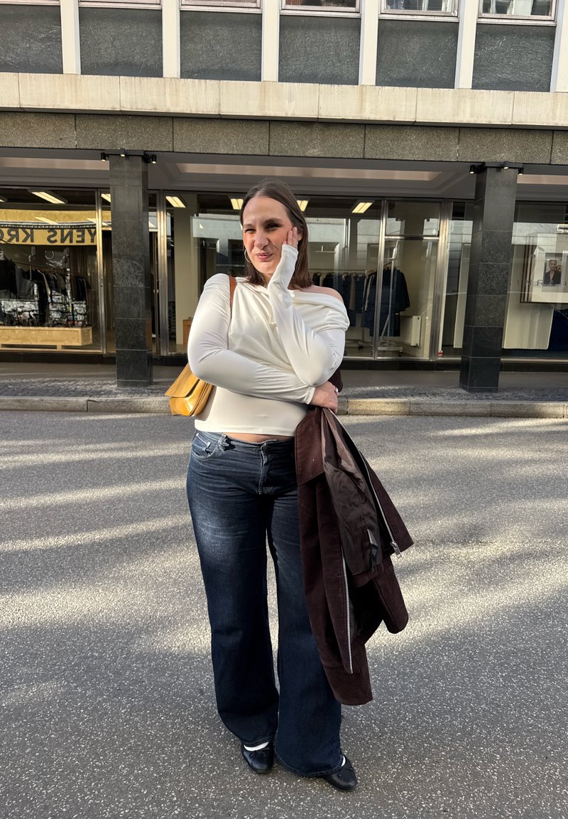 Frau lächelt im Freien in weißem Langarmoberteil und dunklen Jeans, hält eine braune Jacke und eine gelbe Tasche, steht auf einer Straße vor einem Geschäft.