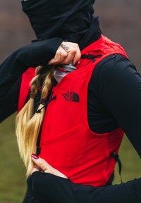 Personne en manches longues noires mettant une bouteille d'eau dans la poche d'un gilet The North Face rouge vif en extérieur.