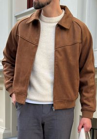 Chaqueta bomber de ante sintético marrón con cuello de solapa, que presenta un cierre de cremallera y detalles de costura sutiles, combinada con un suéter color crema.
