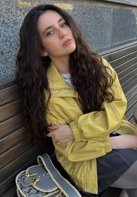 Gelbe Zip-up-Jacke mit elastischen Bündchen, kombiniert mit einem dunkelgrauen Rock. Eine gemusterte Handtasche mit goldenen Akzenten liegt in der Nähe auf einer Holzbank.