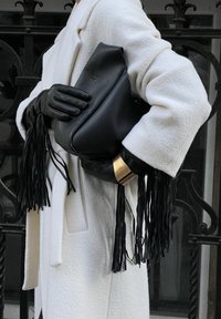 Μαύρη δερμάτινη τσάντα χειρός με φούντες τοποθετημένη απέναντι από ένα λευκό υφασμάτινο παλτό. Οραστά είναι και μαύρα δερμάτινα γάντια καθώς και ένας χρυσός βραχιόλι.