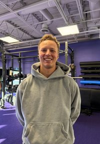 Sweat à capuche gris avec une poche avant, porté par-dessus un T-shirt ; debout dans une salle de sport avec un sol violet et du matériel d'exercice en arrière-plan.