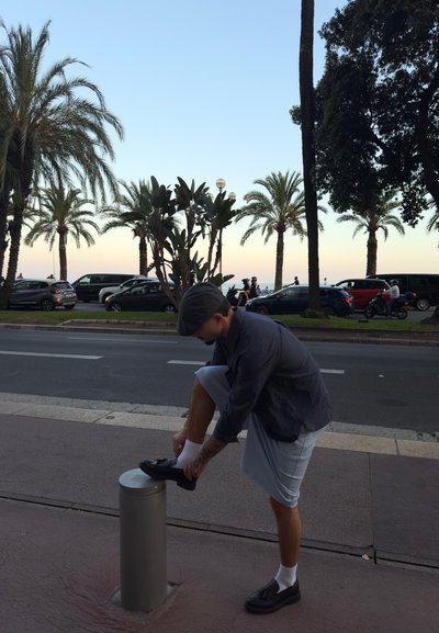 Hombre ajustando un zapato negro sobre un poste de concreto, vistiendo una camiseta a rayas y pantalones cortos de color azul claro, con palmeras y coches de fondo.