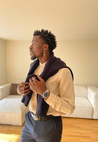 Bărbat cu păr scurt și creț purtând o cămașă bej și un pulover bleumarin așezat peste umeri într-un living luminos.