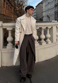 Long manteau blanc, pull en maille crème, pantalon large foncé, et chaussures noires. Debout contre une balustrade en pierre décorative dans un cadre urbain.
