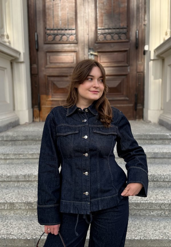 Femme portant une veste en denim foncé et un pantalon se tenant sur des marches en pierre devant de grandes portes doubles en bois, regardant sur le côté en souriant.