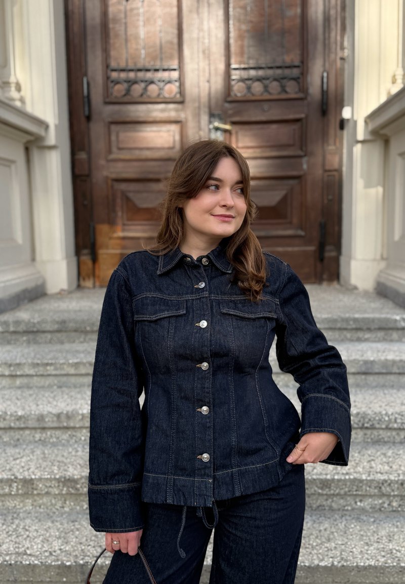 Femme portant une veste en denim foncé et un pantalon se tient sur des marches en pierre devant de grandes portes doubles en bois, regardant sur le côté avec un sourire.