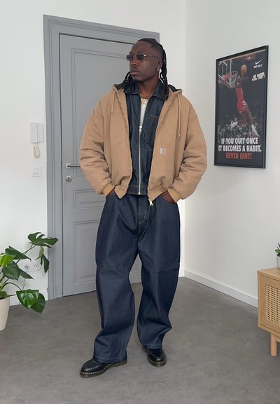 Hombre con chaqueta color caqui, conjunto de mezclilla oscura y gafas de sol, de pie en el interior cerca de una puerta gris y un póster motivacional de baloncesto en la pared.