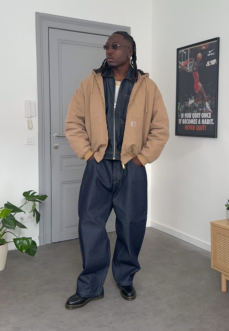 Man wearing tan jacket, dark denim outfit, and sunglasses stands indoors near a gray door and motivational basketball poster on wall.