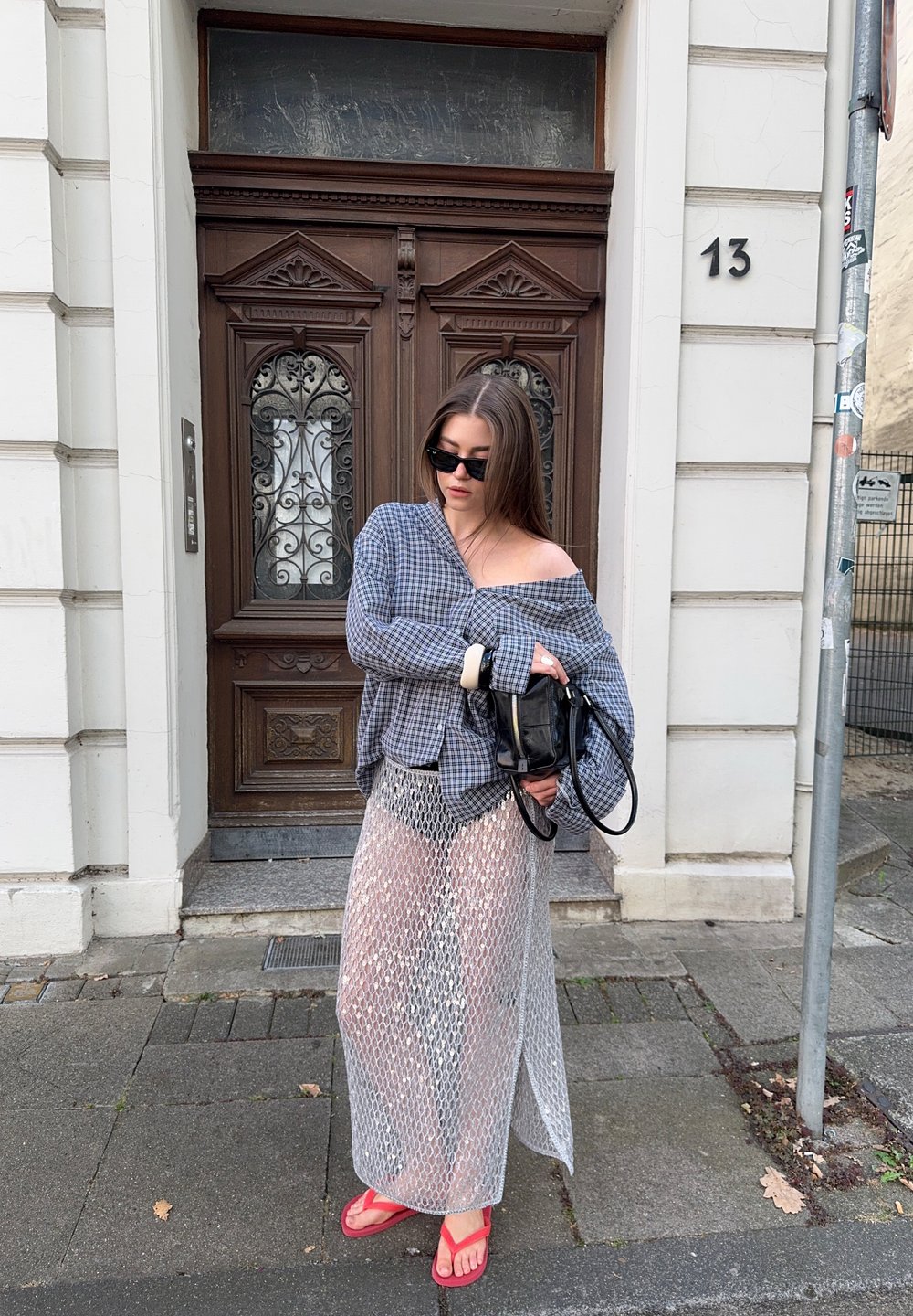Mujer con una camisa de cuadros oversize, una falda larga de malla transparente, chanclas rojas y gafas de sol, sosteniendo un bolso negro cerca de una puerta de madera.