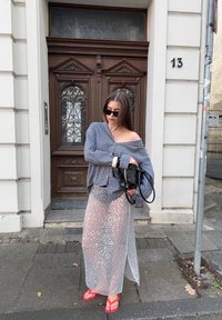 Mujer con una camisa de cuadros oversize, una falda larga de malla transparente, chanclas rojas y gafas de sol, sosteniendo un bolso negro cerca de una puerta de madera.