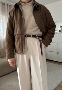 Veste marron courte portée sur un pull gris clair, associée à un pantalon beige taille haute et une ceinture marron. Tissu texturé et coupe décontractée.