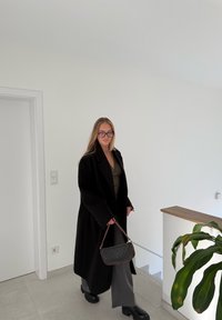 Lungo cappotto nero, top verde oliva, pantaloni grigi a gamba larga e stivali neri con zeppa, che tiene in mano una borsa marrone con motivo decorativo. Ambiente interno luminoso.