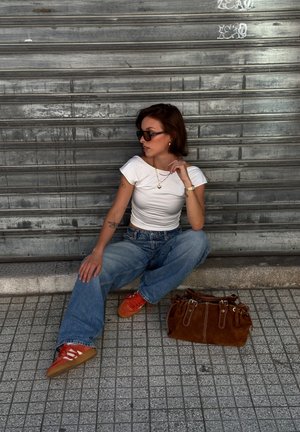 White fitted t-shirt, high-waisted blue jeans, orange sneakers with white accents, and a brown suede handbag. Urban background.