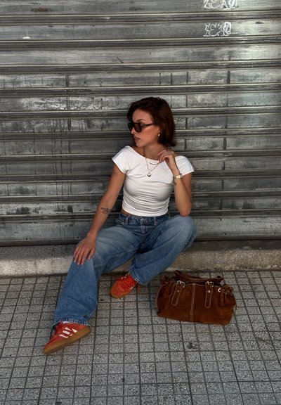 Camiseta blanca ajustada, jeans azules de cintura alta, zapatillas naranjas con detalles en blanco y un bolso de ante marrón. Fondo urbano.