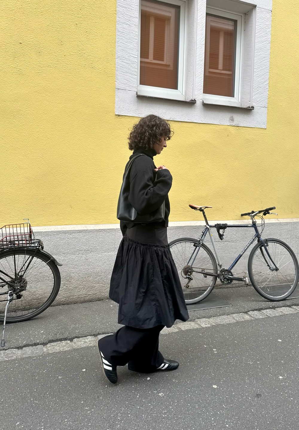 Fekete pulóver, rétegezett fekete szoknya és fekete cipők fehér csíkokkal. Biciklik parkolnak a közelben egy szürke utcán, sárga fal előtt.