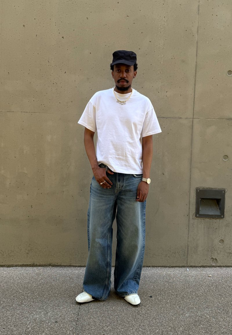 Man som bär svart keps, vit t-shirt, vida blå jeans, vita skor, guldkedja och klocka står mot en beige betongvägg.