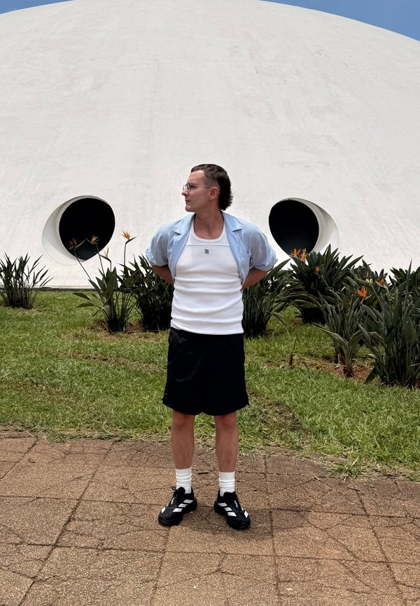 Mann mit Brille, weißem Tanktop, blauem Hemd und schwarzen Shorts steht mit den Händen hinter dem Rücken vor einer weißen Kuppel mit runden Fenstern und Pflanzen.