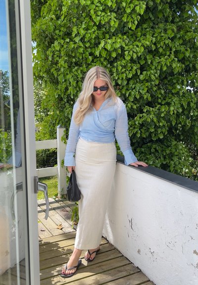 Camisa de manga larga azul claro con tejido texturizado, combinada con una falda de lino cruda de talle alto. Bolsa negra y chanclas. Fondo de vegetación verde.