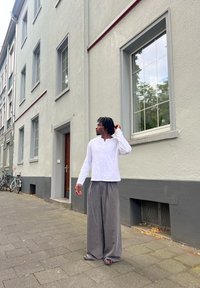 Camisa branca de mangas longas e calças largas cinzas em uma pessoa, em pé na calçada ao lado de um edifício cinza com várias janelas e uma porta de madeira.