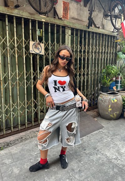 Joven con camiseta sin mangas "I love NY", pantalones cortos grises rotos, calcetines rojos, zapatos negros y gafas de sol, de pie en un callejón urbano con plantas y bicicletas.
