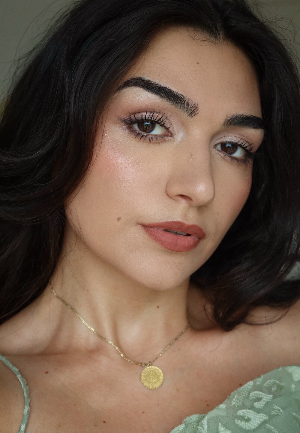 Collana d'oro con un ciondolo circolare, labbra marrone chiaro e tonalità della pelle soffice. Capelli scuri e ondulati, con una maglietta verde a fantasia floreale.