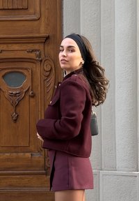 Jachetă scurtă burgundă cu butoane decorative, asortată cu o fustă mini. Păr lung brunet cu o bentiță neagră, ținând o poșetă mică neagră.