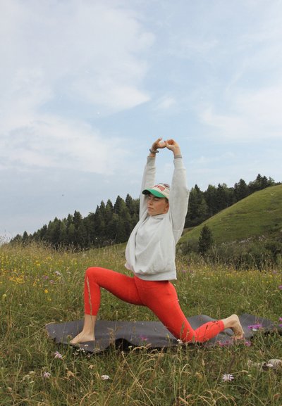 Una mujer con un sudadera gris claro y leggings rojos realiza una postura de yoga sobre una esterilla oscura, rodeada de flores silvestres y colinas verdes.